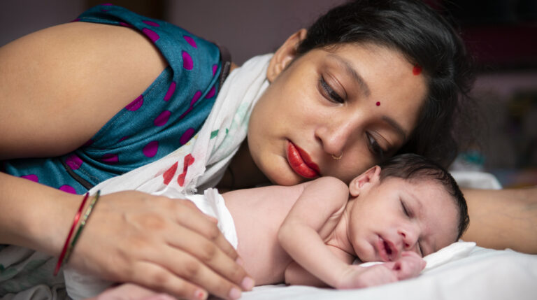 A mother in a sari tenderly cradles her newborn baby, both of them lying on a bed, in an intimate, peaceful scene.